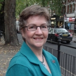 Photo of Joanna Biddolph, wearing a bright teal blue jacket, standing on Chiswick High Road.