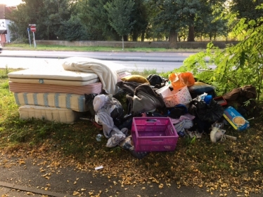 Photo of mattresses, soiled clothing, sacks and general waste piled on a grass verge beside a road